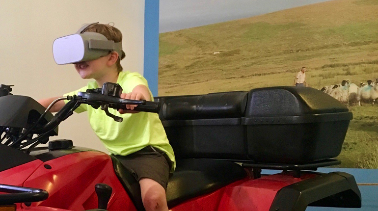 Child on quad bike wearing a virtual reality headset, with photo of sheep on Dartmoor in the background