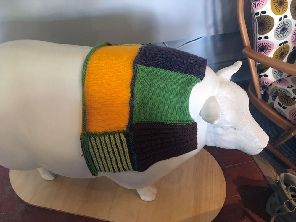 model sheep wearing part finished patchwork knitted coat in green and yellow squares knitted by the MEWES knitters 
