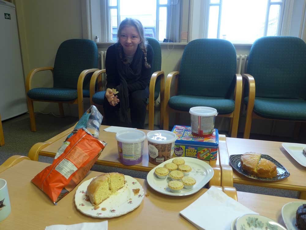 Student sitting in the staff room behind a table full of cakes and snacks before preparing her Arts Award exhibition for Takeover Day