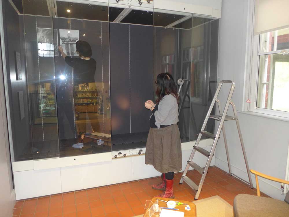 Student standing inside a big glass case putting up a picture with a member of staff looking on