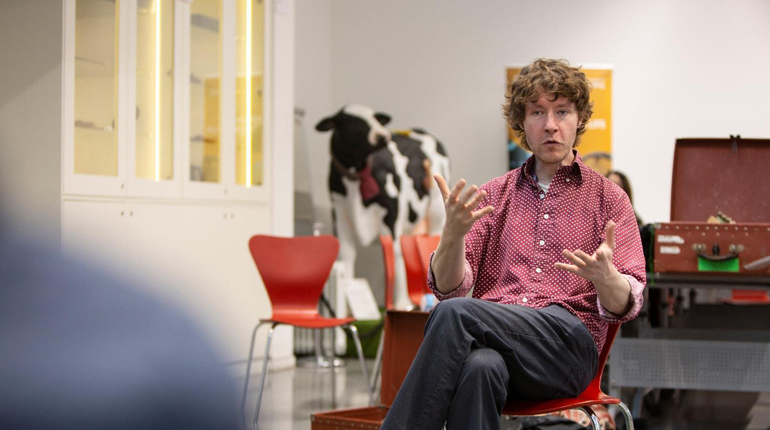 Pete Flood sitting and gesturing whilst speaking with MERL cow in background
