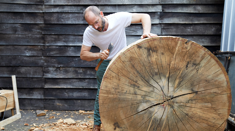 Maker Gareth Neal standing behind large flat circle of wood with an axe working on Shoulder to the Wheel exhibition piece