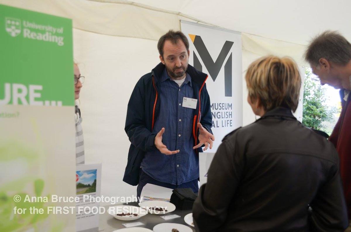 A man standing in a tent and behind a table covered with bowls of dried insects, speaks to people on the other side of the table