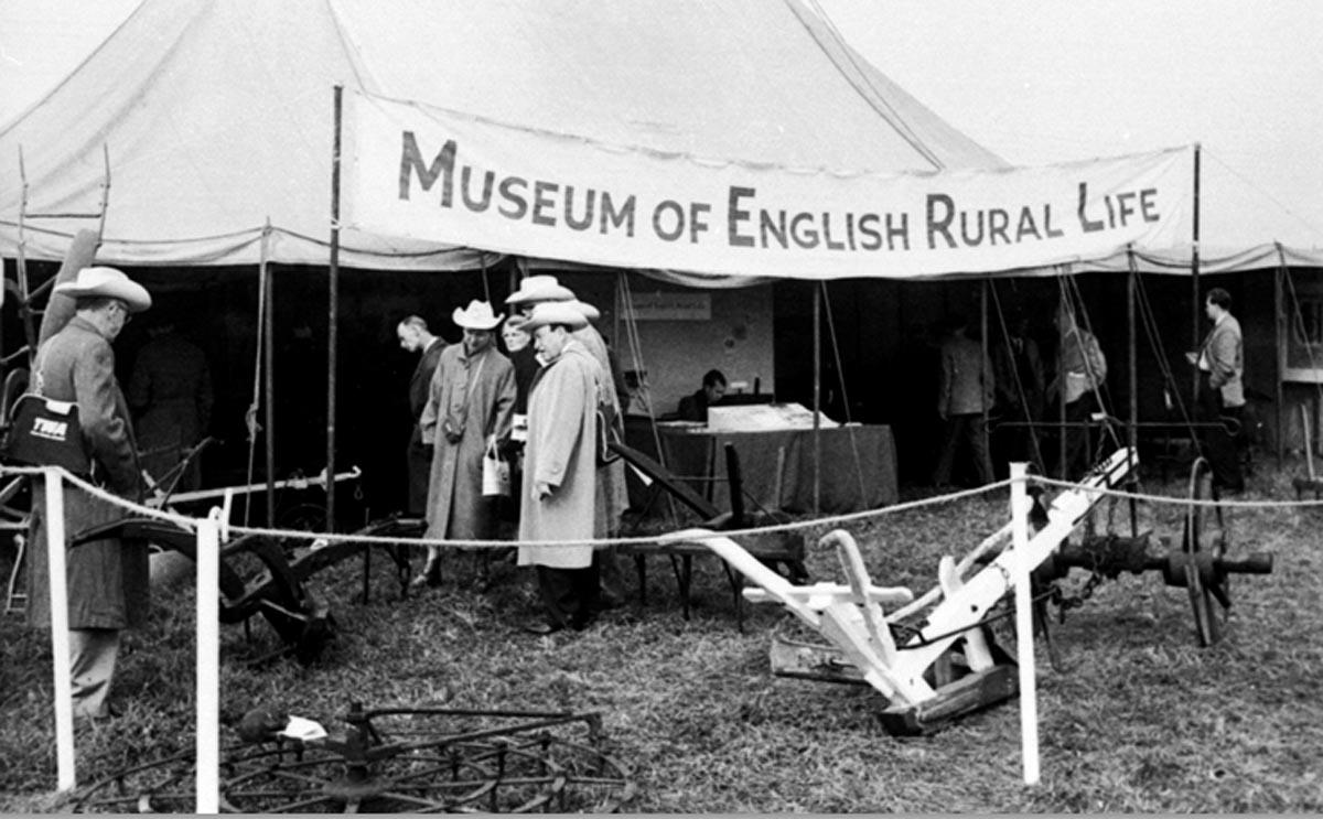 Men in cattlemen's hats congregate outside a tent with a sign reading 'Museum of English Rural Life', and farming implements displayed prominently in front.