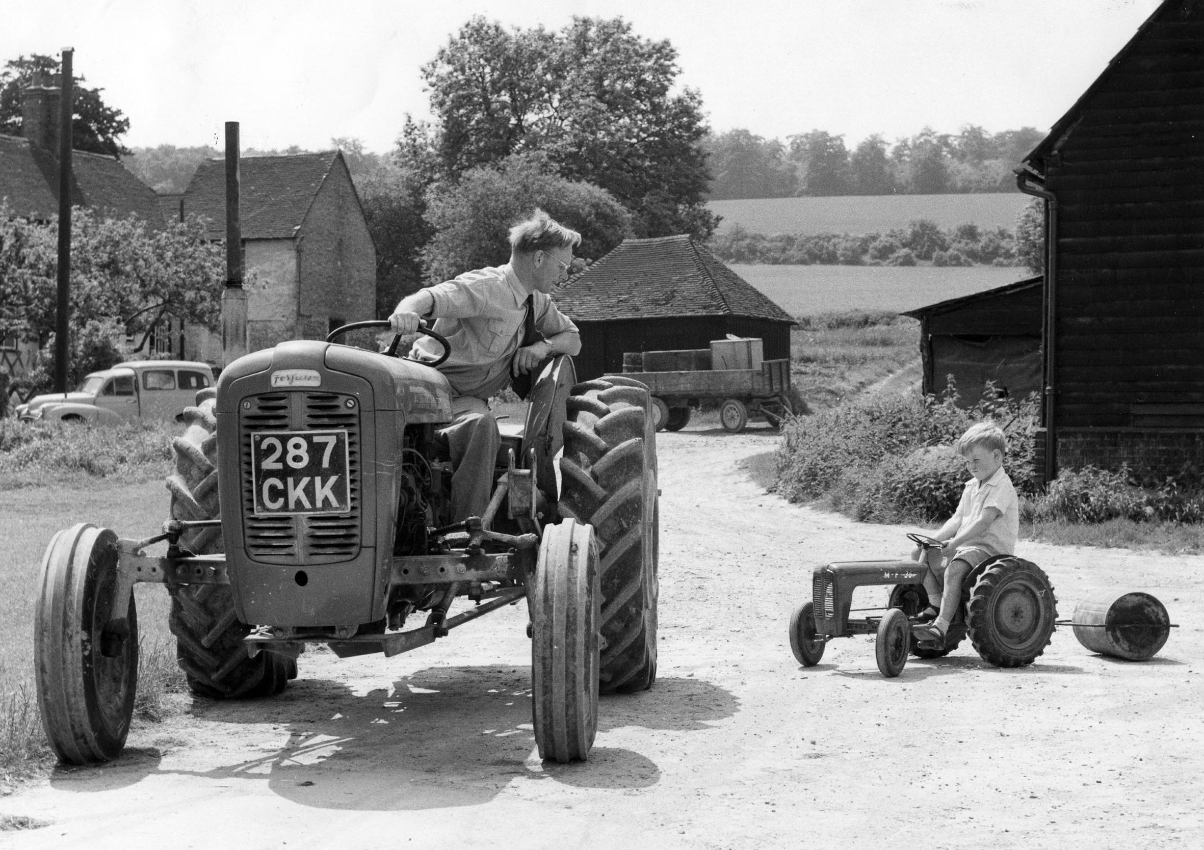 Man sitting on a tractor looks down to boy sitting on pedal tractor, with farmyard and countryside in the background