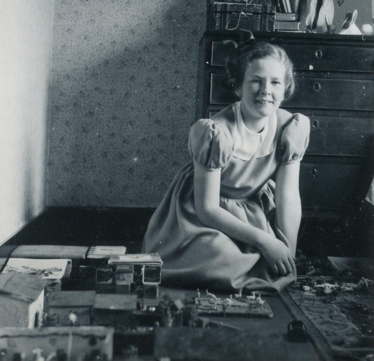 Black and white image of a young girl sitting on the floor of a bedroom with homemade farm buildings in front of her and farm toys