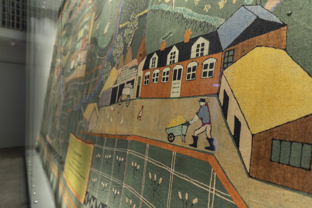 Detail of stylized textile design showing figure with wheelbarrow, farmyard, and fields. Blurred edge of display case appears in background indicating that this is on display in a gallery space.