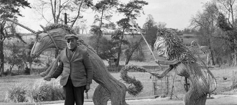 Black and white photograph of a man with an eye patch and cap standing beside large straw sculptures of a lion and a unicorn, with trees in the background.