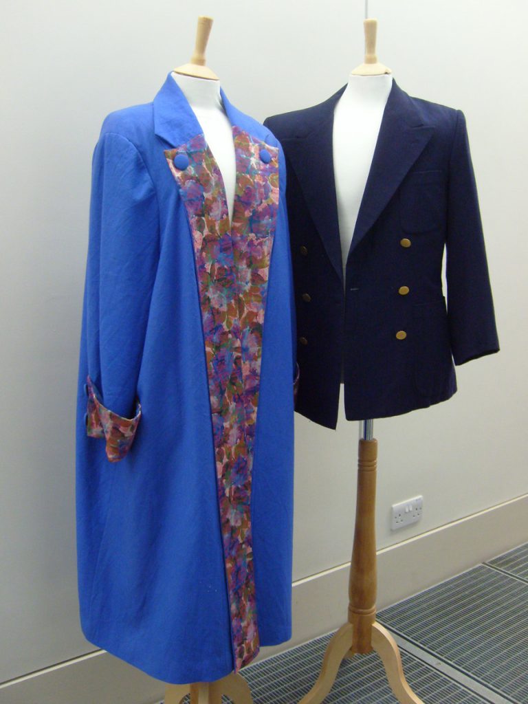 On the left a bright blue ladies coat with a multicoloured detail on the cuff and lapels. On the right a short navy blue men's blazer with three gold buttons. Both are displayed on mannequins.
