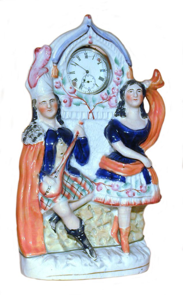A painted pocket watch holder. It is made from pottery and features two Highland dancers. The dancers have deep blue outfits on with a kilt on one dancer and a floral skirt on the other.