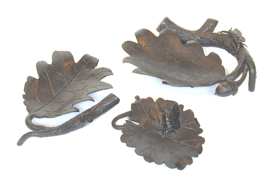 Three iron ornaments. from left to right a plain leaf, a leaf with a small butterfly on it and a leaf attached to a twig with an acorn and an insect.