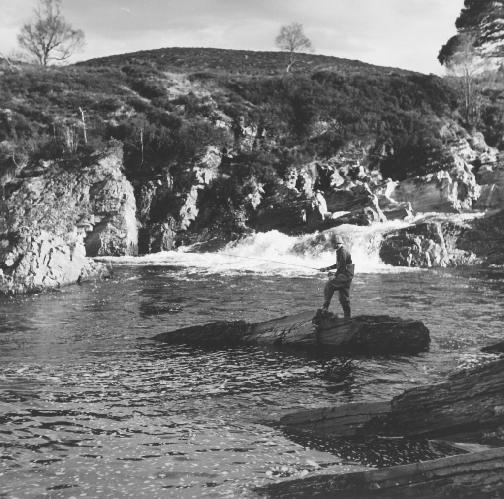 A black and white image of a man flyfishing on a river. He is standing on an island of rocks in the middle of the river.