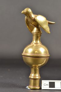 A brass polehead of the bedpost type with a single bulge, and surmounted by a small bird with downward pointing wings.