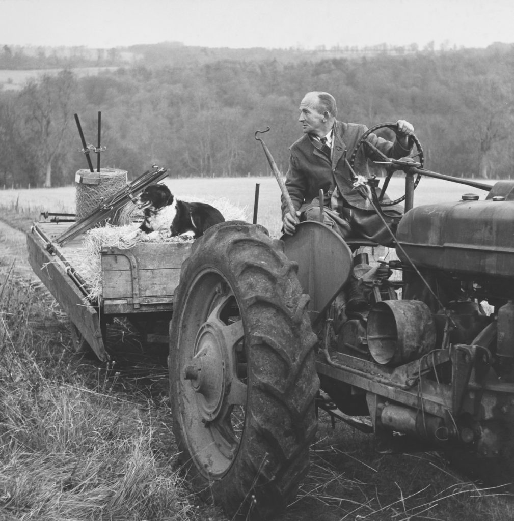 A black and white photograph of a farmer in a tractor pulling a trailer with a sheepdog on it. He may be one of the secretive Tractor Whisperers.