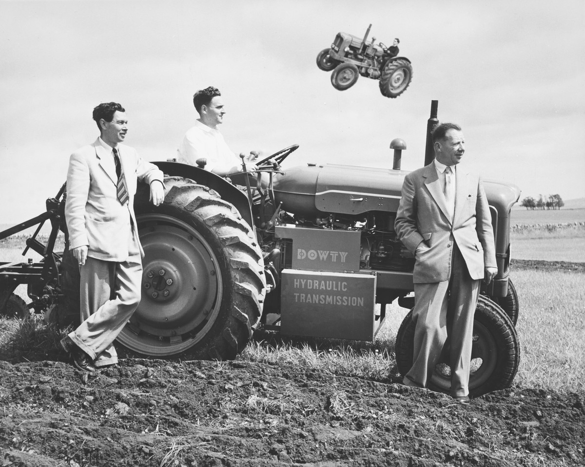 Blank and white photograph of 3 suited men with a Dowty tractor. Behind them another tractor soars majestically through the clouds. Very likely Tractor Whisperers.