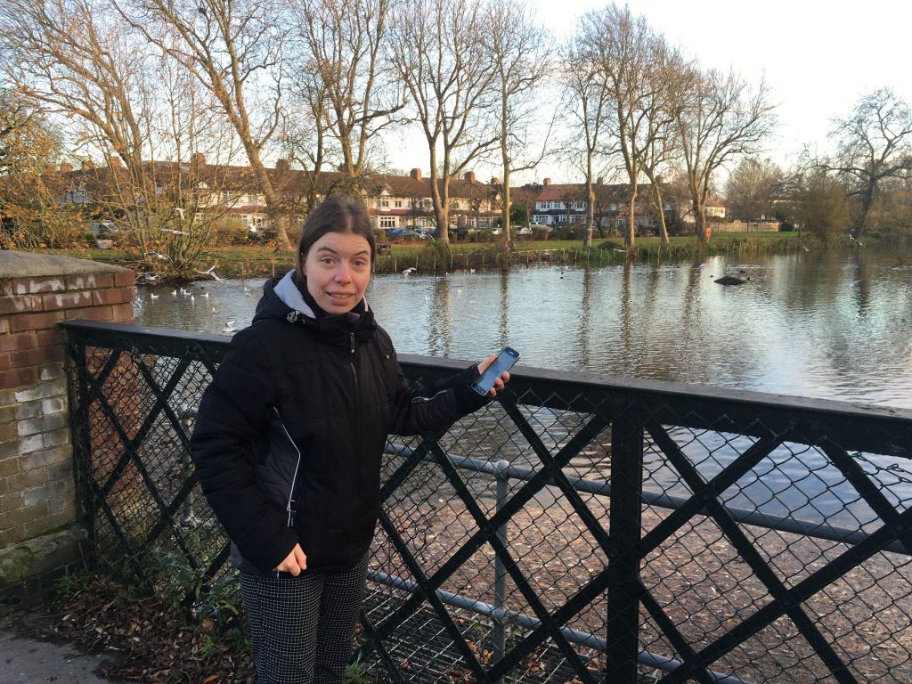 Woman standing holding a mobile phone on a bridge over a pond. There are trees and houses in the background.