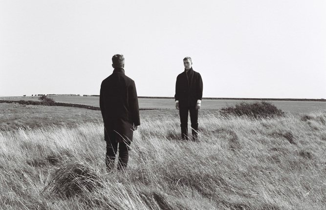 A black and white photograph of two people in the middle of a field, facing each other.