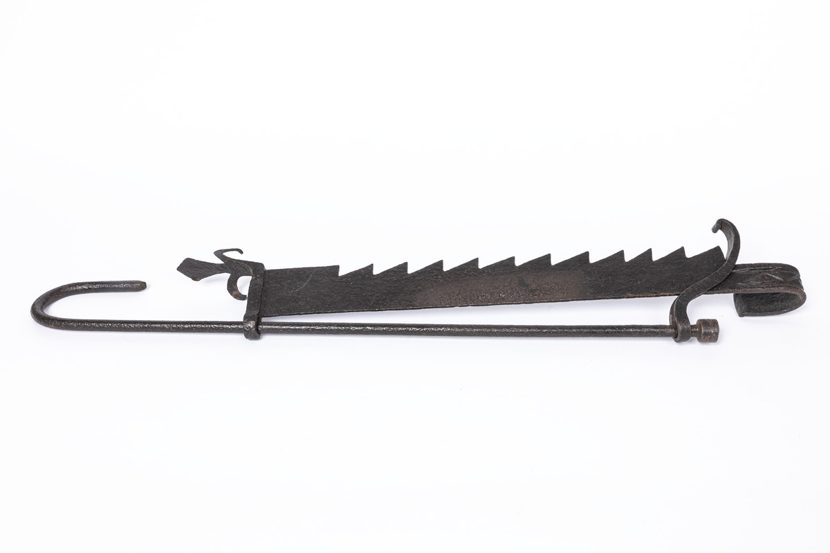 This is an eighteenth-century Sussex-made wrought iron ratchet hanger, used for suspending cooking utensils over an open hearth. It consists of a toothed rack with a hook at one end and a <em>fleur-de-lys</em> ornamentation at the other. The ratchet is attached by a shackle to the hooked suspension rod. (59/252)