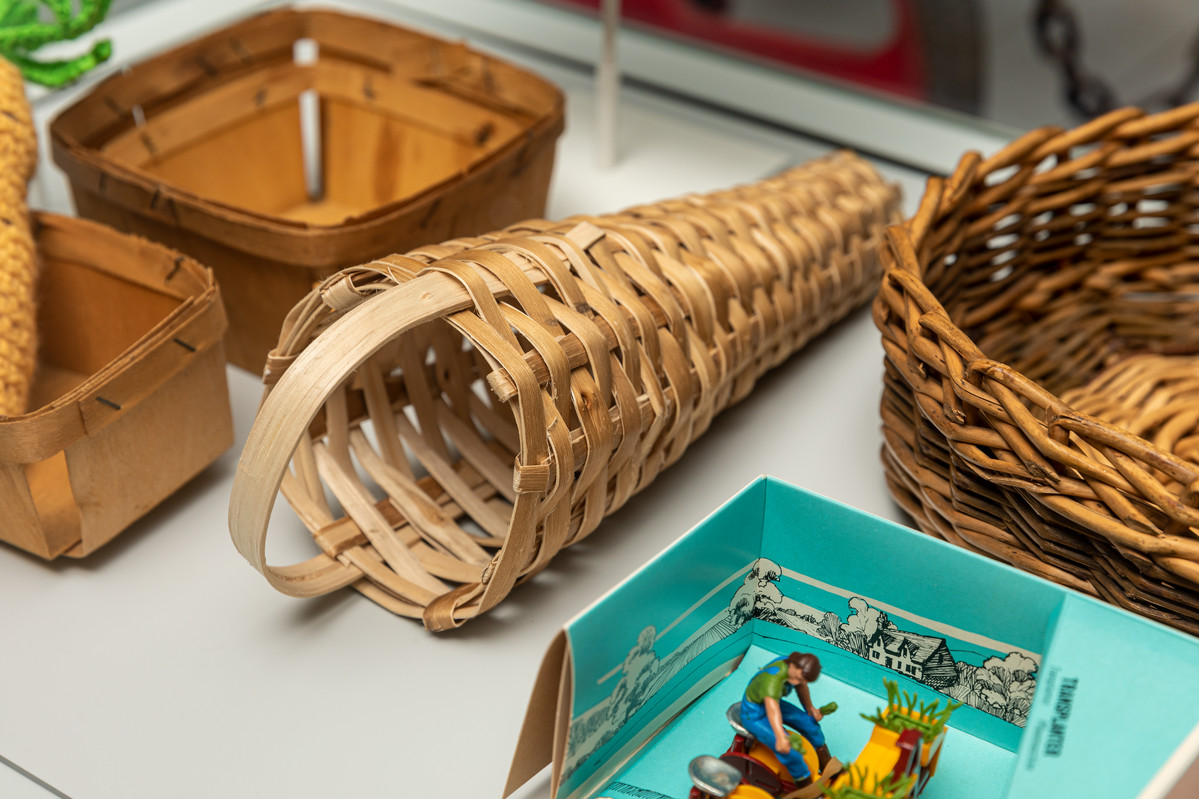 This image shows a long thin basket used for soft fruits, displayed alongside other objects in a museum display case.