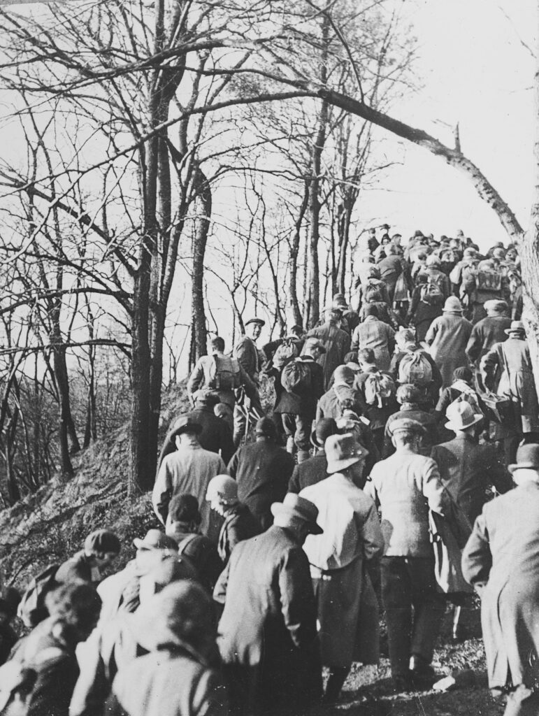 A large group of men and women in hiking clothes and with their backs to the camera hike up the ridge of a hill.