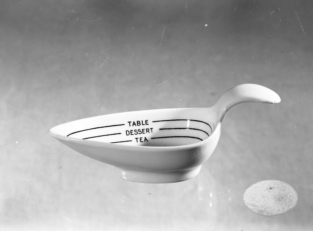 This is a porcelain measuring spoon used when cooking or for measuring medicine. It is white, with 'tea', 'dessert' and 'table' marked around the inside.