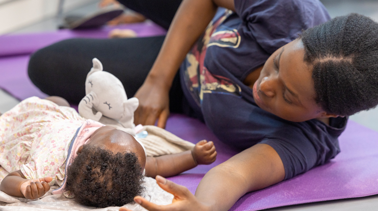 A mum lies next to a baby on a mat as they take part in a Mums and Baby yoga session at The MERL