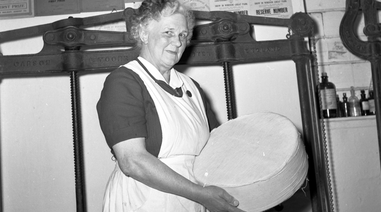 A black and white photo of lady in an apron holding a large round of double Gloucester cheese
