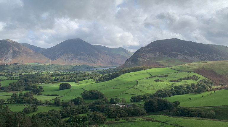A photograph of the a Lake District landscape with green fields and trees in the foreground and fells in the distance