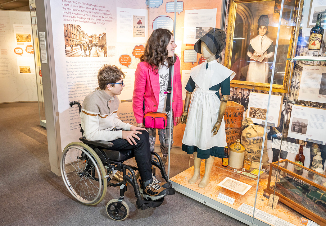 Young people in the galleries at Reading Museum. One of them is sitting in a wheel chair and the other is standing as they both look at a case containing a mannequin