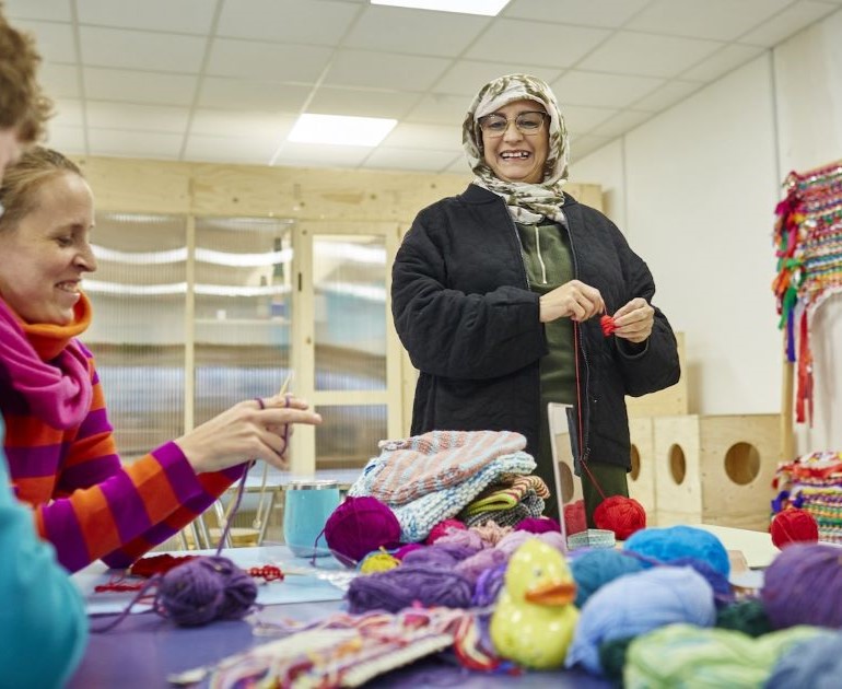 Women smiling and knitting together; standing and sitting at a table filled with colourful balls of wool.