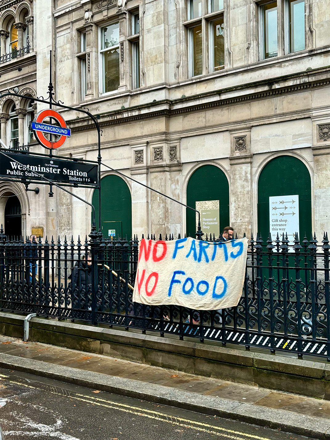 A protest banner hangs over a London underground station exit. It reads: 'No Farms, No Food'.